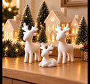 Kit 3 renas de Natal decorativas, cor Branco, em frente a uma vila de Natal iluminada